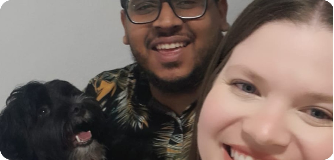 Casal sorrindo para a foto. O homem segura um cachorro preto de pelos compridos que o observa enquanto a foto é tirada