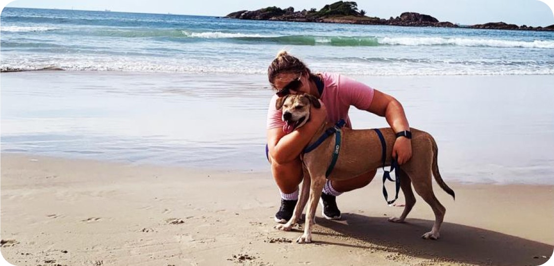 Mulher abraça e beija um cachorro caramelo na areia da praia, com a vista do mar ao fundo