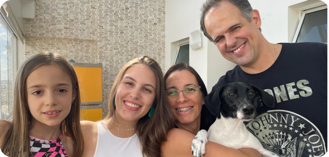 Quatro pessoas de uma família sorriem para foto, onde a mulher segura um cachorro preto e branco no colo