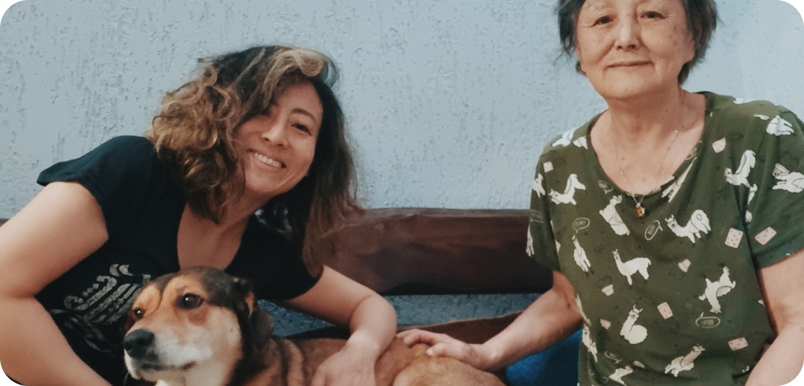Cachorro caramelo, preto e branco, sentado entre duas mulheres que olham sorrindo para a foto