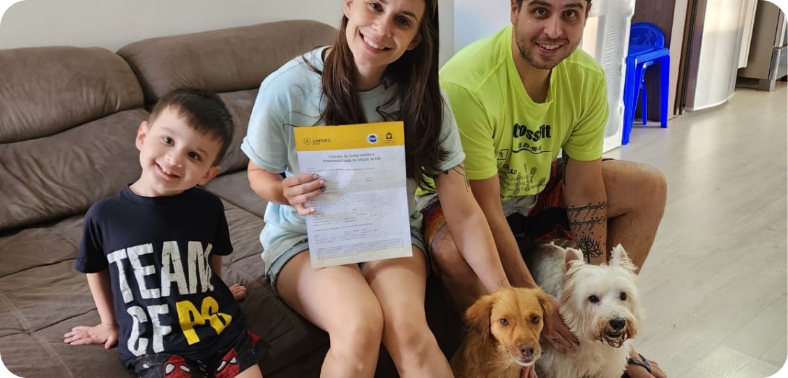 Mulher sentada expondo um termo de adoção em uma das mãos. Em um de seus lados está um homem e um menino do outro lado, todos sorrindo com dois cachorros sentados entre eles.