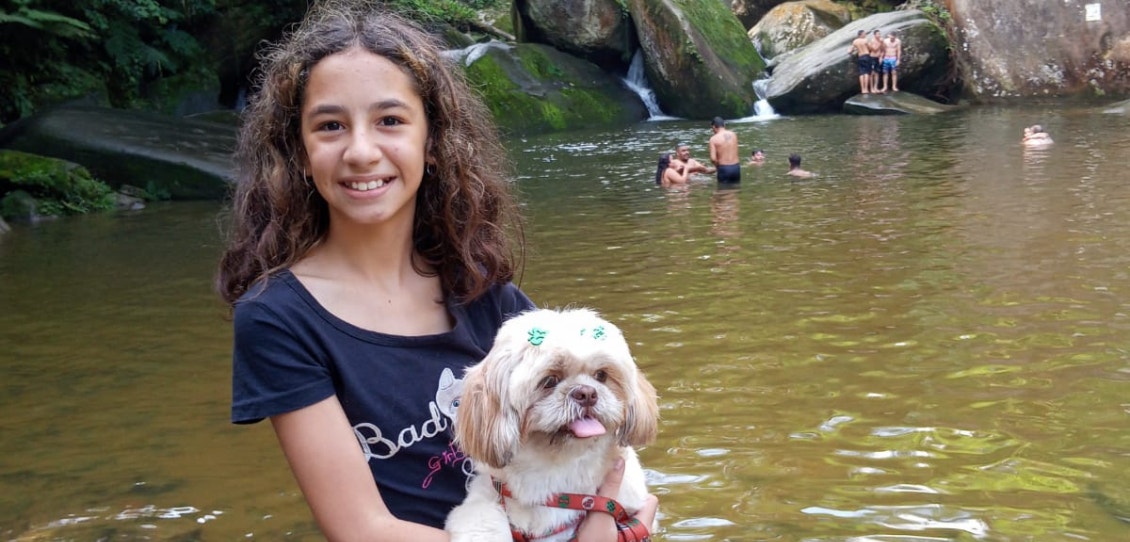 Menina segurando uma cachorrinha peluda caramelo e branco com um lago e pedras ao fundo.