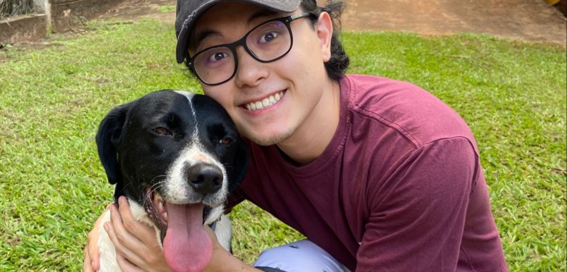 Homem de óculos, boné e camisa vinho, abraçado a um cachorro preto e branco. No fundo, gramado e árvores.