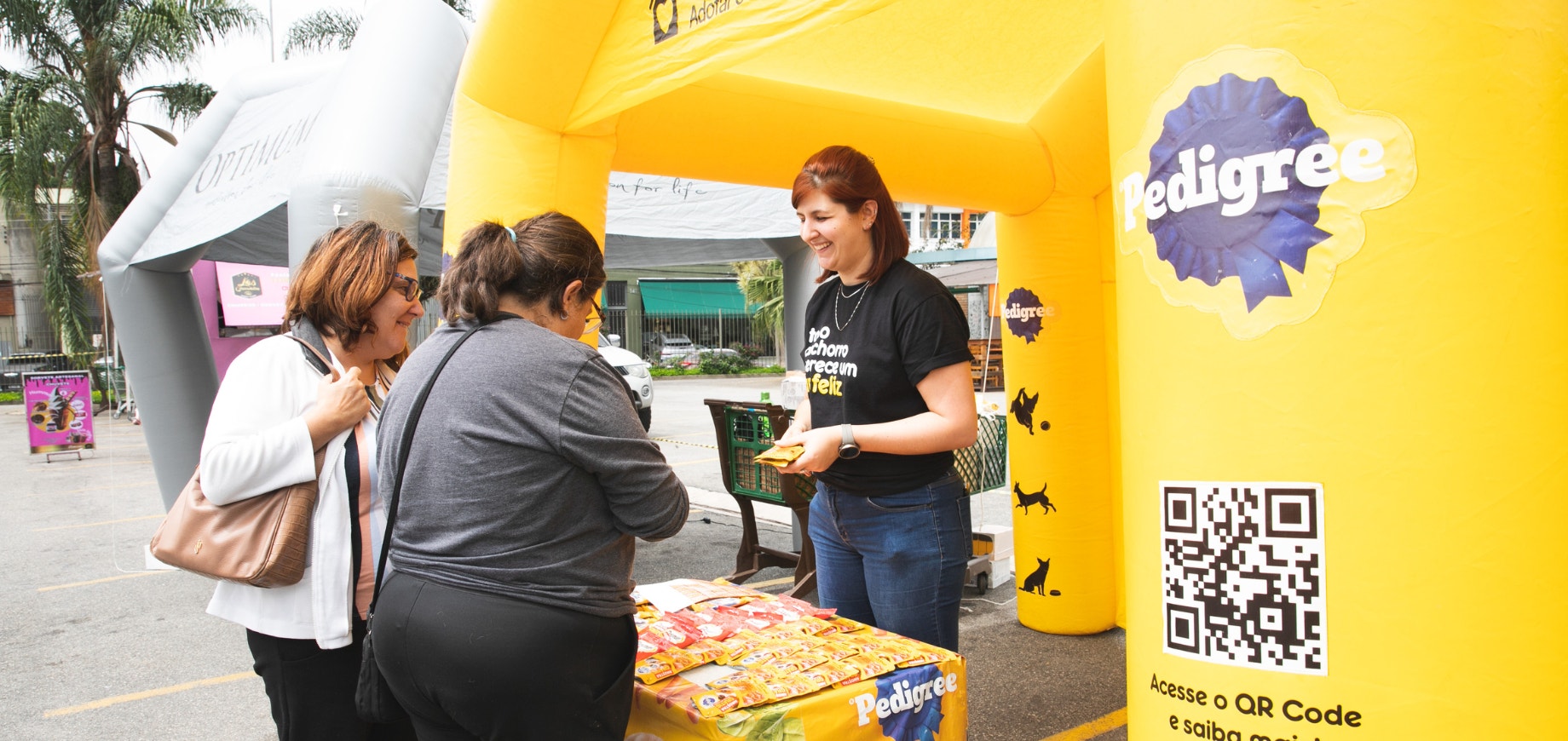 Tenda inflável amarela com logo da PEDIGREE, com uma moça dentro atendendo duas mulheres que escutam mais sobre o programa Adotar é tudo de bom.
