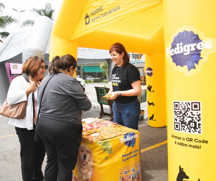 Tenda inflável amarela com logo da PEDIGREE, com uma moça dentro atendendo duas mulheres que escutam mais sobre o programa Adotar é tudo de bom.