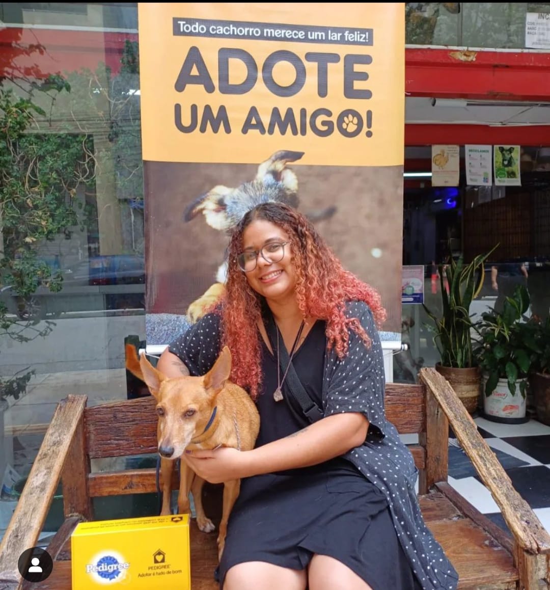 Fotografia da Bárbara sentada em um banco de madeira ao lado do seu cachorro Bob com uma placa 'adota um amigo' ao fundo'.