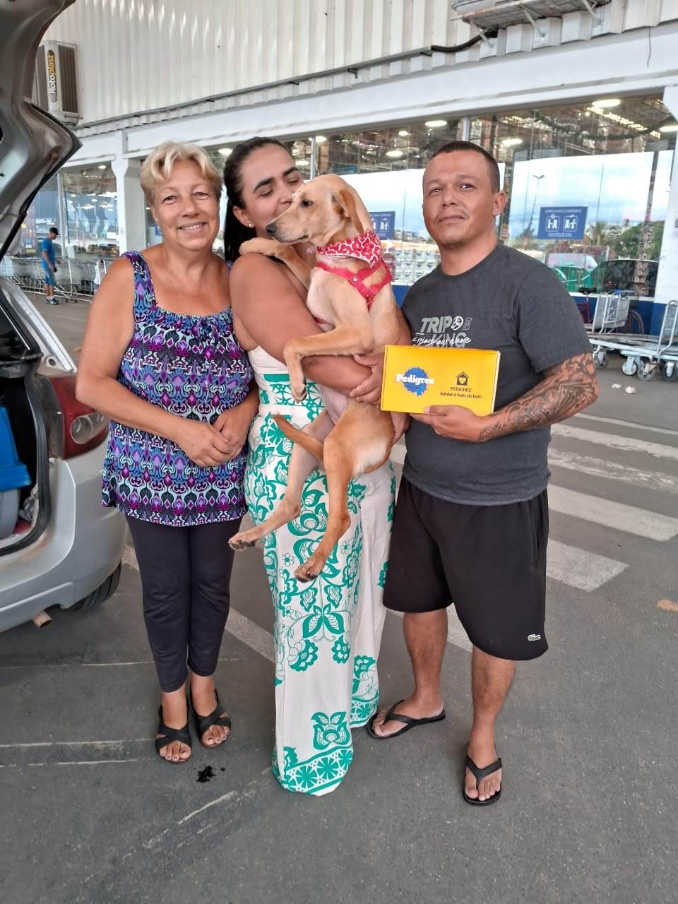 A imagem mostra três pessoas sorridentes com o cão Laura com pelugem bege em um estacionamento. A mulher do centro segura Laura no colo e beija sua cabeça, enquanto a mulher à esquerda e o homem à direita, que segura uma caixa de ração Pedigree, olham para a câmera. Ao fundo, há um prédio com janelas de vidro, sugerindo ser um supermercado ou loja de departamentos.