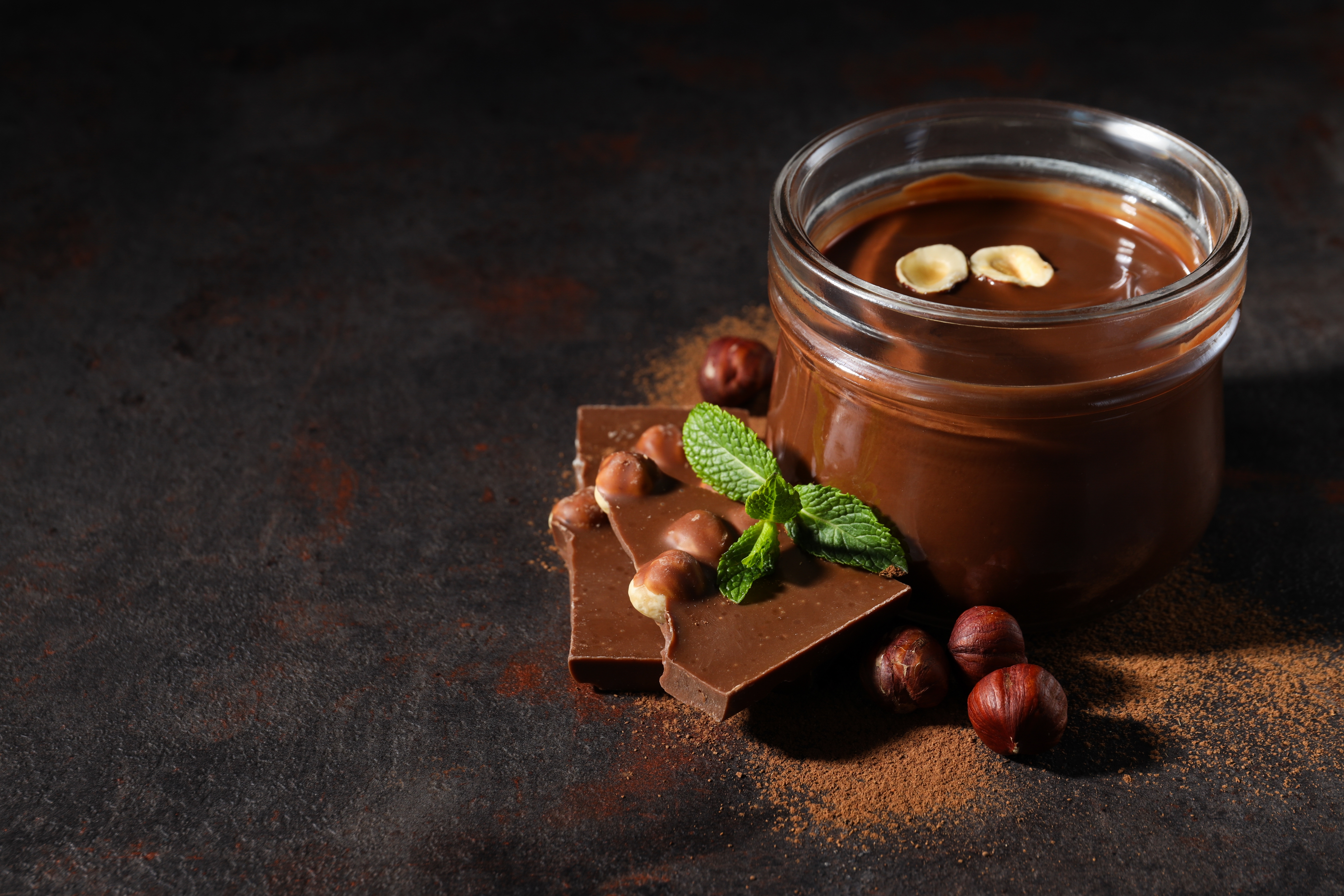 Pote de creme de avelã com pedaços de chocolate, avelãs inteiras e folhas de hortelã sobre uma superfície escura polvilhada com cacau em pó.